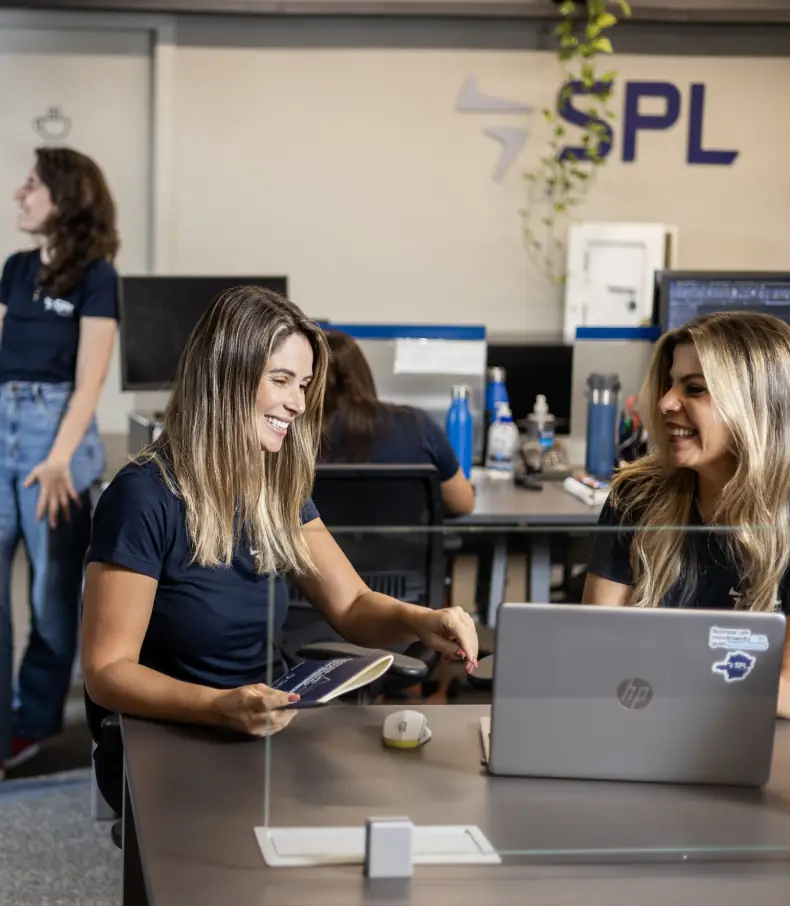 spl slide 3 Duas colaboradoras sorriem e interagem em frente a um notebook em uma mesa de escritório da SPL, com outras pessoas e a logo da empresa ao fundo.