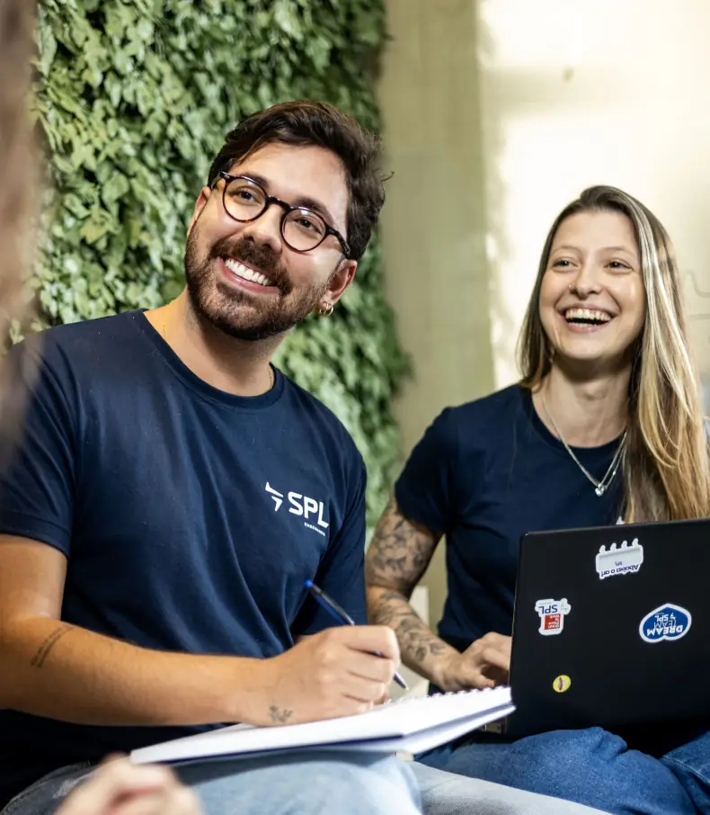 spl slide 4 Um colaborador de óculos sorri e anota em um caderno ao lado de uma colega de trabalho também sorridente, ambos vestindo a camiseta da SPL em um ambiente de escritório com uma parede verde ao fundo.